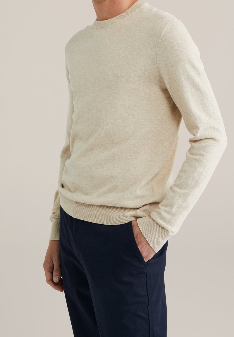 Homme portant un pull beige à manches longues et un pantalon bleu marine foncé, une main dans la poche, sur un fond uni.
