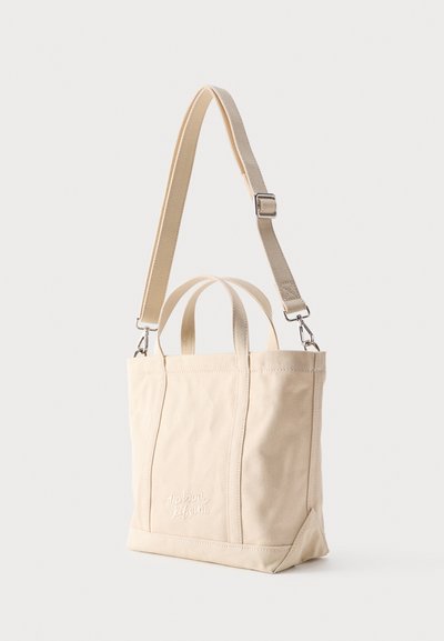 Μπεζ καμβά τσάντα ώμου με δύο κοντές λαβές, έναν ρυθμιζόμενο μακρύ ιμάντα και την κεντημένη επιγραφή «Maison Kitsuné» στο μπροστινό μέρος.