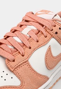 Nike sneaker featuring a white leather base, peach suede accents, textured laces, and perforated detailing on the toe area.
