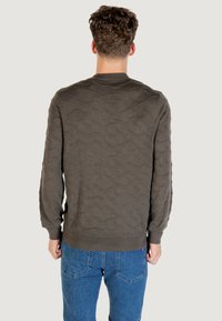 Olivgrüner strukturierter Pullover mit einem wellenförmigen Muster, gerippten Bündchen und Saum. Über blauen Jeans getragen, die eine anliegende Passform zeigen.