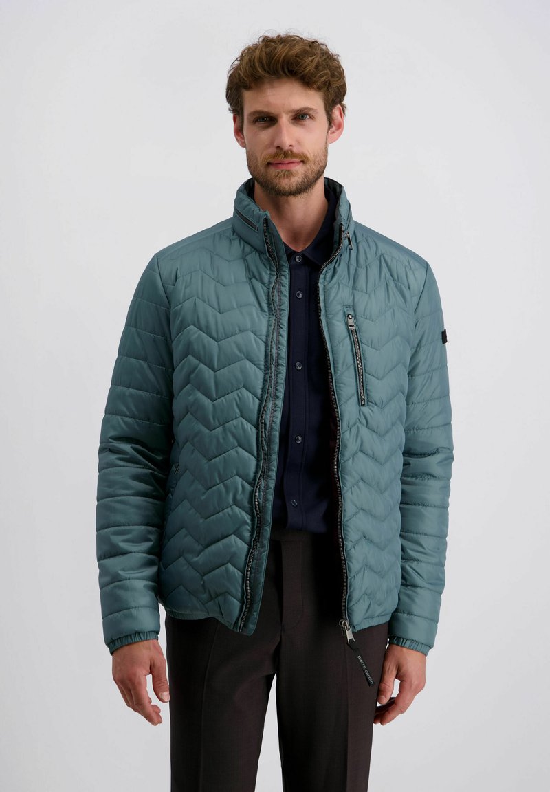 Man in een teal gewatteerde jas over een donker knoopshirt en donkere broek, staande tegen een effen lichte achtergrond.