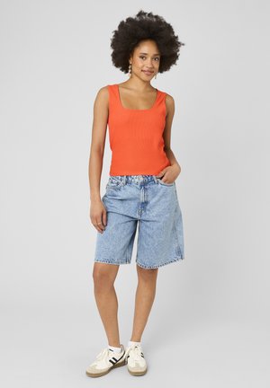 Femme avec coiffure afro portant un haut sans manches orange, un short en jean bleu clair jusqu’aux genoux, des baskets blanches et des boucles d’oreilles dorées, debout de façon décontractée.