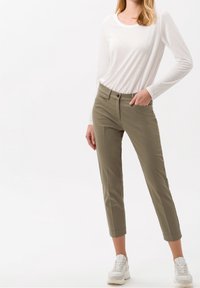 Kobieta ubrana w białą bluzkę z długim rękawem, oliwkowe spodnie 3/4 i białe trampki, z jedną ręką w kieszeni, na białym tle.