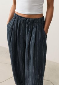 Femme portant un pantalon large bleu marine à fines rayures avec taille à cordon et un débardeur court blanc, mains dans les poches.