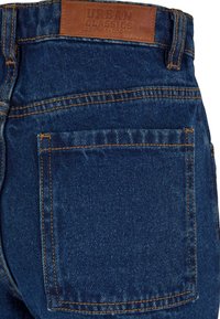 Vue de dos d'un jean en denim bleu montrant une poche et un patch en cuir marron portant l'inscription « Urban Classics Denim Factory ».