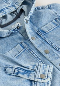 Veste en denim bleu clair avec des boutons-pression en métal, poches poitrine visibles et coutures détaillées sur les manches et le col.