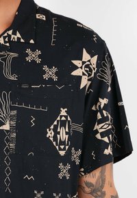 Chemise à manches courtes en tissu noir avec des motifs géométriques et abstraits beiges, comportant une poche poitrine et un col boutonné.
