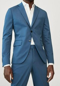 Uomo in abito blu su misura con giacca a due bottoni e camicia bianca, in piedi davanti a uno sfondo chiaro.