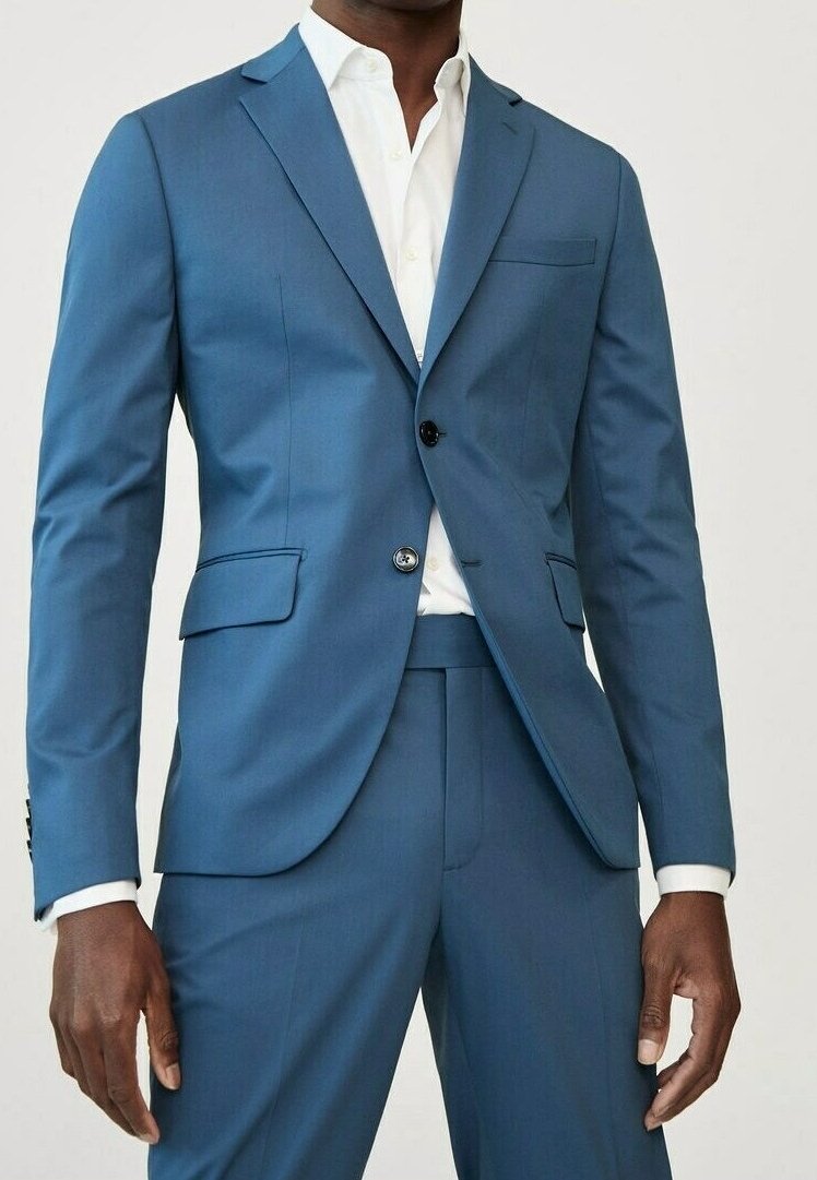 Uomo in abito blu su misura con giacca a due bottoni e camicia bianca, in piedi davanti a uno sfondo chiaro.