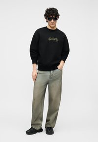 Man with curly hair wearing black graphic sweatshirt, baggy faded gray jeans, black sneakers, and large dark sunglasses, standing relaxed.