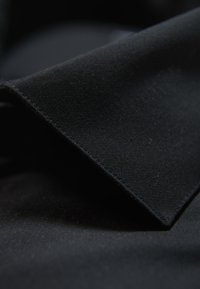 Zwarte stof, strak geweven met een gladde textuur. Het materiaal vormt scherpe, gean angleerde plooien en toont een matte afwerking zonder patronen of accenten.