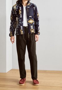 Fleecová bunda s geometrickými vzory v tmavě modré, zlaté a krémové barvě, s kapsou na zip. Kombinována s olivově zelenými kalhotami a červenými botami.