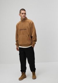 Brązowy polarowy hoodie z pół zamkiem błyskawicznym i napisem "COALITION", z przednią kieszenią, w połączeniu z czarnymi spodniami cargo i brązowymi butami na sznurówki.