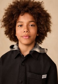 Camicia nera con bottoni, fodera del cappuccio a quadri e tasca applicata. Tessuto dalla texture liscia, con colletto prominente e dettagli in metallo a contrasto.