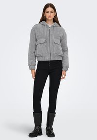 Halli korduroost bomberjakk kapuutsiga, millel on kaks eesmist taskut ja elastsed mansetid, kombineeritud mustade kõrge vöökohaga teksade ja mustade saabastega.