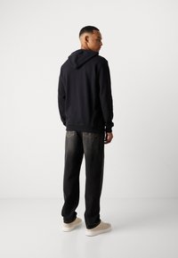 Mann in schwarzem Kapuzenpullover und ausgebleichten schwarzen Jeans steht mit dem Rücken zur Kamera und schaut zur Seite, trägt beige Sneaker vor einem weißen Hintergrund.