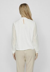 Femme avec de longs cheveux blonds portant un chemisier blanc texturé à manches longues et un pantalon beige, vue de dos sur un fond uni.