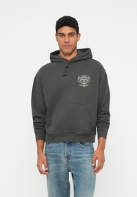 Egy férfi sötétszürke kapucnis pulóvert visel, amelynek a mellkasán egy kis címerlogó található, világoskék farmernadrággal, egy sima fehér háttér előtt áll.