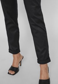 Černé kalhoty na míru s ohrnutými nohavicemi, vyrobené z hladkého materiálu. V kombinaci s otevřenou černou koženou obuví na podpatku se štíhlým stiletto podpatkem.