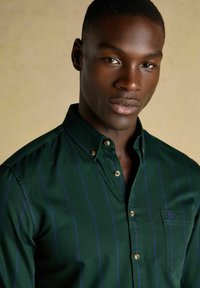 Junger Mann in einem dunkelgrünen Hemd mit dünnen lila Streifen, der vor einem beigen Hintergrund steht und in die Kamera schaut.