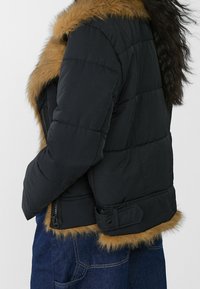 Zwarte pufferjas met een bruine nepbontafwerking, voorzien van een ritssluiting en een taille met ceintuurdetail. Genaaid panelenontwerp zichtbaar.