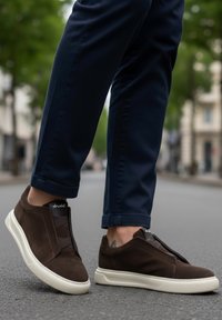Brown suede slip-on sneakers with a textured upper, elastic detailing, and a white rubber sole, paired with navy trousers.