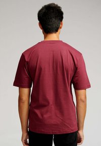 Mand med kort sort hår står med ryggen til iført en ensfarvet maroon kortærmet t-shirt og sorte bukser foran en neutral baggrund.