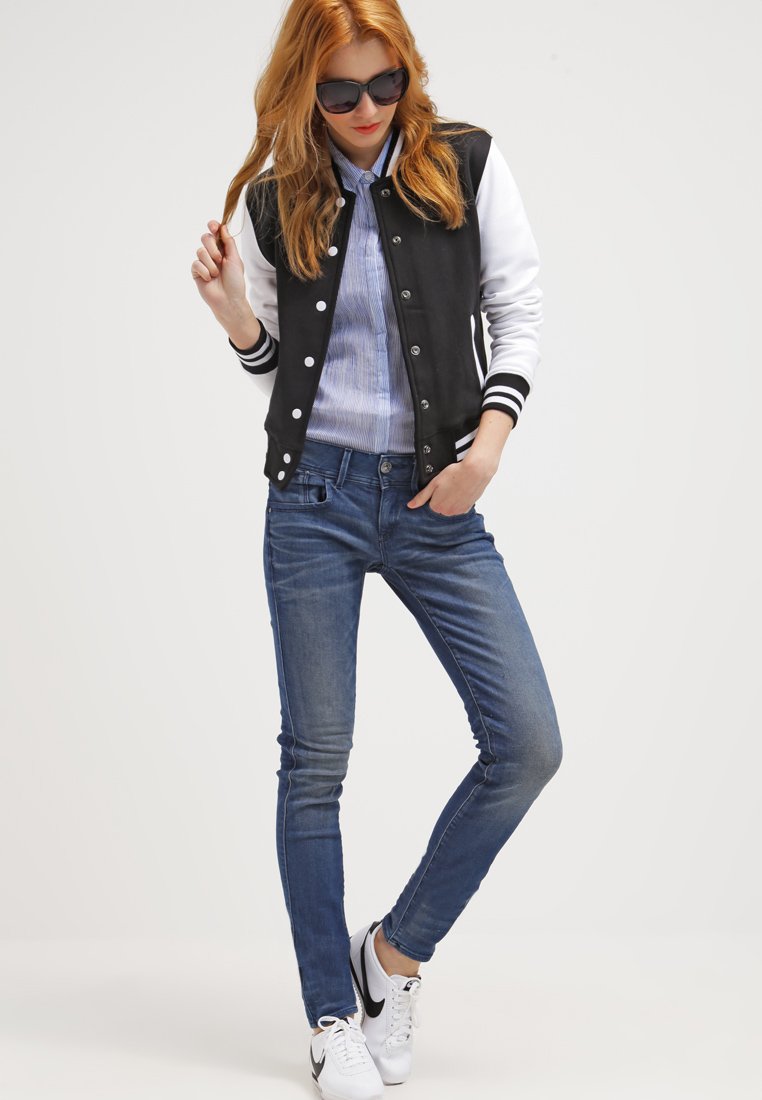 Chaqueta varsity en blanco y negro con puños a rayas, camisa de botones azul, jeans ajustados azules y zapatillas blancas con acentos negros.