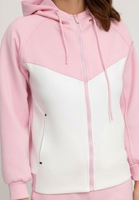 Sweatshirt à capuche zippé avec des panneaux roses et blancs, tissu doux, poignets côtelés, et deux poches latérales avec des accents noirs.