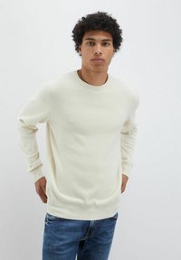 Maglione lavorato a maglia di colore crema con scollatura tonda, tessuto testurizzato, polsini e orlo a costine; abbinato a jeans di denim blu.