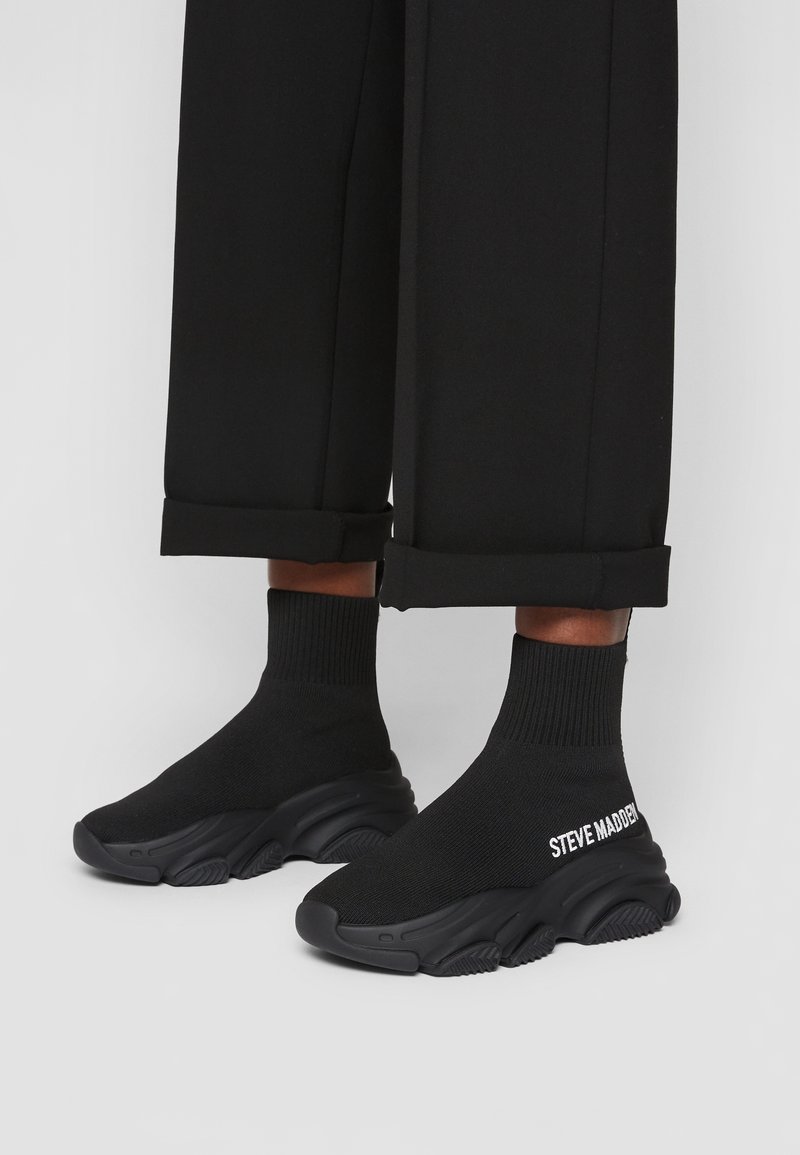 Baskets style chaussettes noires avec semelles épaisses et texte « STEVE MADDEN » sur le côté, portées avec un pantalon noir retroussé sur un fond clair.