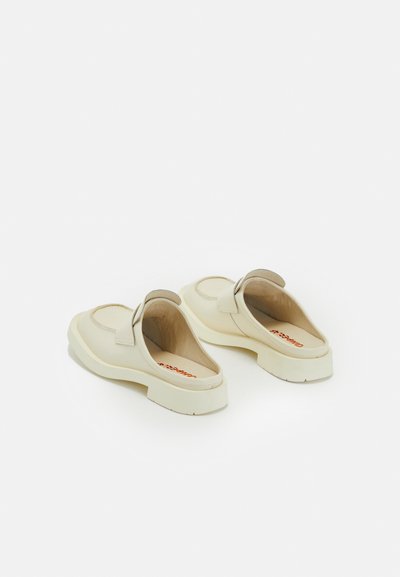 Krämsfärgad slip-on-skor med rund tå, mjukt ovandel och tjock strukturär sula. Har en loopad detalj vid hälen och en synlig varumärkeslogotyp i rött.