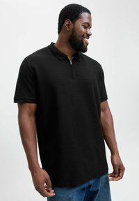 Sort polo-shirt med lynlås foran, korte ærmer og en tekstureret stof. Har et lille logo på brystet og en afslappet pasform.