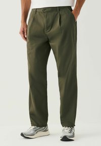 Khaki green pants with a straight fit, pleats at the front, and side pockets. Paired with gray sneakers featuring a mesh design.