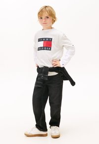 Grå långärmad T-shirt med en svart och röd rektangulär logotyp, kombinerad med svarta jeans och vita sneakers. Denimjacka knuten i midjan.