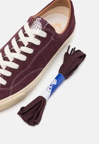 Burgundy suede sneaker with beige laces, featuring a textured rubber sole. Includes a matching burgundy shoelace and a blue label.