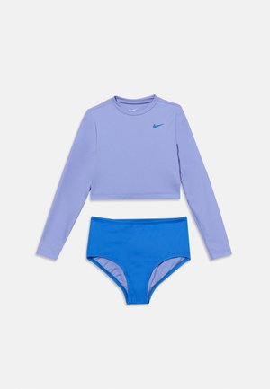 Top corto de manga larga en color lavanda con el logo Nike en azul, combinado con bragas de baño de talle alto en azul sobre fondo blanco.