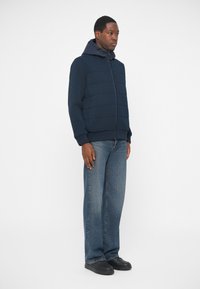 Ναυτικές νάιλον μπουφάν με φερμουάρ, υφή πάνελ και κουκούλα; συνδυασμένα με ανοιχτού μπλε παντελόνια με φαρδιές γραμμές και μαύρα παπούτσια.