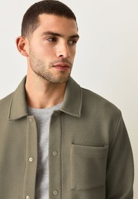 Giacca verde oliva con un motivo testurizzato, dotata di colletto a camicia, chiusura a bottoni a pressione e una tasca sul petto sopra una camicia grigia.