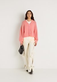 Ροζ κοντό κασκόλ με μακριά μανίκια και κουμπιά, σε συνδυασμό με κρεμ παντελόνι που έχει λεπτομέρειες από φιόγκο στο στρίφωμα και μαύρη τσάντα με πουά σχέδιο.