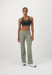 Mujer atlética con trenzas, vestida con un sujetador deportivo negro, pantalones de yoga verde oliva y zapatillas blancas, de pie frente a un fondo liso.