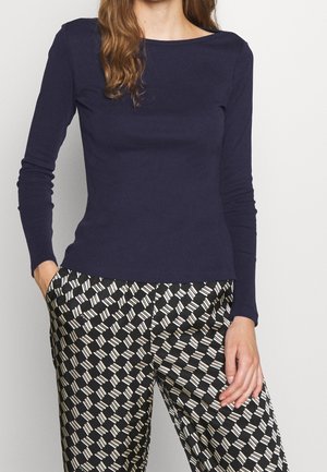 Kobieta ubrana w dopasowany granatowy longsleeve i czarne spodnie o szerokich nogawkach z białym geometrycznym wzorem, stojąca z jedną ręką w kieszeni.