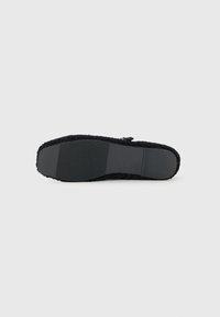 Suela plana negra de un zapato de tipo slip-on con fondo de goma texturizada y bordes forrados de fleece, fotografiada sobre un fondo blanco liso.