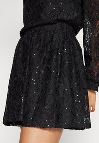 Svart spetsklänning med paljetter, med en utsvängd kjol. Strukturerat blommönster, långa ärmar och detalj med elastisk midja.
