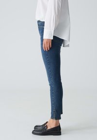 Jean skinny bleu foncé avec un léger stretch, présentant une taille mi-haute et des détails de couture subtils, associés à des mocassins noirs.
