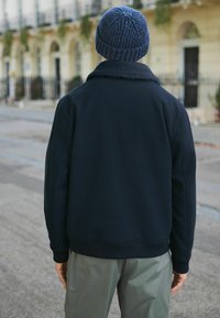 Henkilö, jolla on päällään tumma takki ja sininen neulottu pipo, katsomassa poispäin, seisomassa ulkona kaupungin kadulla, taustalla rakennuksia.