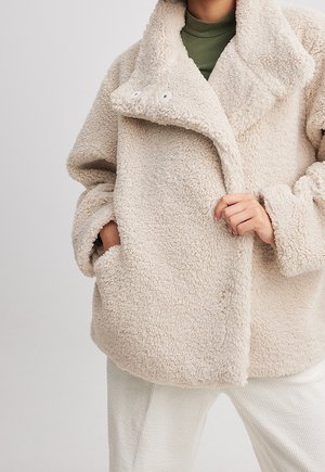 Personne portant une veste sherpa beige et duveteuse avec un grand col, un col roulé vert et un pantalon blanc, main dans la poche de la veste.