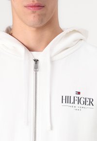 Valge tõmblukuga kapuutsiga jakk, millel on kangast tekstuur, esiküljel "HILFIGER" logo ning rinnal väike punane ja sinine lipu embleem.