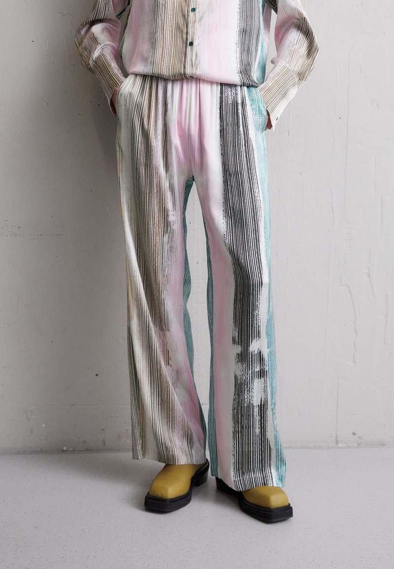 Pruhovaný shirt a široké kalhoty v pastelových barvách, včetně růžové, tyrkysové a hnědé. Hladká látka s jemnou texturou. Žluté dřeváky jsou součástí.