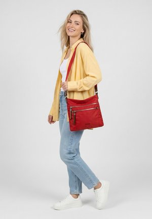 Vrouw met een geel shirt, wit topje, lichtblauwe spijkerbroek, witte sneakers, draagt een rode crossbodytas met meerdere ritsvakken.